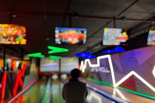 A person, how hands are hidden in front of them, stands just before an open lane at a bowling alley. All pins are set for a fresh take at the bowl. Neon lights are all around, the room feels dark and cool. It is ready to be played.