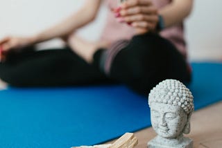 woman in pink top and black leggings sits on blue yoga mat holding burning sage, in the front there is a Buddha statue and bowl of burning Palo Santo