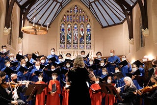 A medium-sized choir of adults and children stands in formation, in choir robes, poised to sing with a chamber orchestra in a church; you see the back of the conductor, poised to conduct.