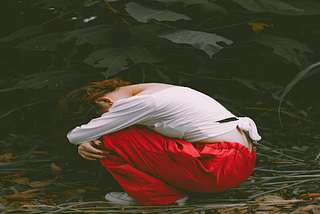 A person wearing a white long-sleeve top and red pants crouches on the ground in a forest, head down and arms wrapped around their knees, surrounded by fallen leaves and dark green foliage, conveying a mood of sadness or introspection.
