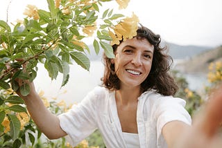 A photo of a woman with dark hair and light brown skin smiling next to a tree with yellow flowers.