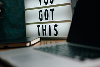 A picture of a small white sign with black letters on a desk next to a laptop. The sign says “YOU GOT THIS”.