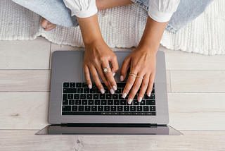 Photo of a seated woman on the floor wearing gold rings, typing on a laptop by Mikhail Nilov via Canva.