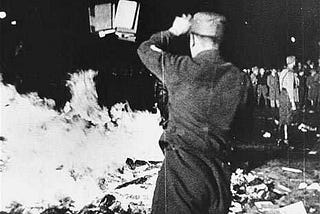 A black and white, grainy photo of a pile of books in the street, on fire. A crowd is visible looking on, and in the foreground, a Nazi soldier facing away from the camera throws several more books onto the pile.