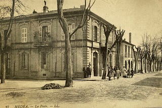 Black and white picture of the post office in Medea, this is a two story European style building on a tree-lined boulevard. A group of people, including men in suits and girls in white dresses is seen posing in front of the building.
