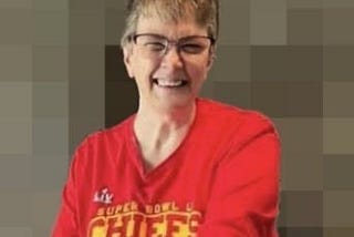 A smiling woman with brown hair with grey in it, wearing glasses and a red Kansas City Chiefs long sleeved shirt.