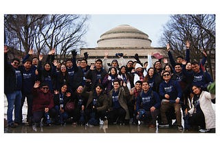 About 50 individuals, most wearing BREIT sweatshirts, wave in a group photo with the MIT Dome behind them