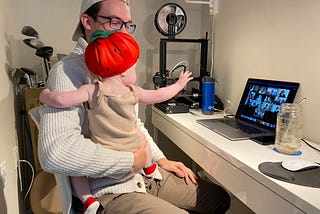 Rylan sits with his 1-year-old daughter, Avery, on his lap while she waves to people visible in a Zoom meeting on Rylan’s laptop.