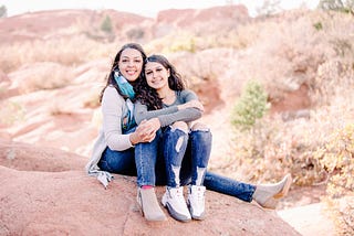 A half-black mother with mid-length dark brown hair, wearing a grey sweater and blue jeans, hugs her teenage daughter, a half-black girl with long brown hair, wearing a long sleeve gray shirt and blue jeans. They are sitting atop a rock at Garden of the Gods park in Colorado.