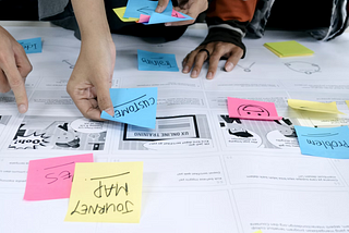 Photo of a table with printed papers, post-it notes and people interacting with the notes in a workshop.