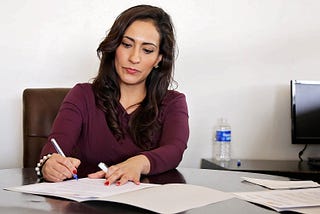 An attractive dark haired lady sits writing at an office desk.