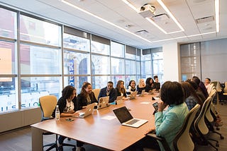 Several women of color and a couple of men sit at a large light brown conference room table with laptops in front of them. The conference room has large windows. The atmosphere appears fun and lighthearted.