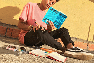 Bruno é um homem negro de cabelos crespos castanhos. Está vestindo uma camiseta rosa, uma calça com rasgos no joelho e um tênis preto. Ele está lendo o livro “Ideias para adiar do fim do mundo”, de Ailton Krenak, que tem uma capa azul. Bruno está sentado no chão, encostado em uma parede amarela com um rodapé de tijolos. No chão, há um estojo com canetas coloridas e um livro aberto.