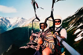 A man and woman are shown in harness gliding. Snow capped mountains are shown in the background