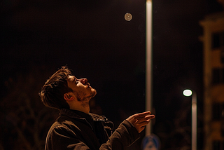 Image of a man standing under a streetlight at night flipping a coin
