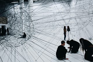 People exploring a large-scale installation visualizing a complex network structure, with black strings forming interconnected nodes in a white room, symbolizing connectivity and interdependence.