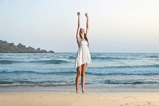 A woman on a beach holds her arms skyward as though manifesting by reaching upward