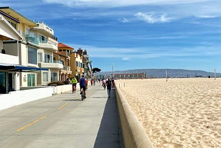 a pedestrian-only street next to the beach, with houses on one side and sand on the other. it’s a blue-sky day.