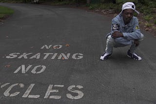 Rapper NGB QUIS squatting down wearing a white hat with a jean jacket