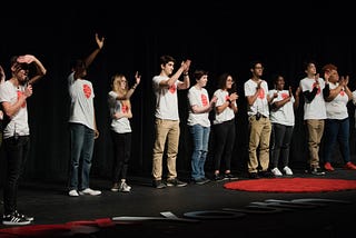 A group of young people, all wearing event-themed shirts, stands on a stage, claps, and smiles.