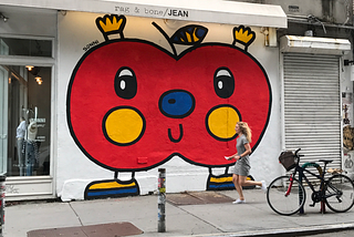 Street art — a red apple by the artist Sonni is raising its arms and rejoicing, on a white wall in NYC Bowery. A white woman in a sporty dress runs past the apple in a hurry.