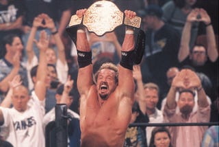 A blond man, Diamond Dallas Page, holds a golden belt over his head. Behind him, a crowd cheers him on.