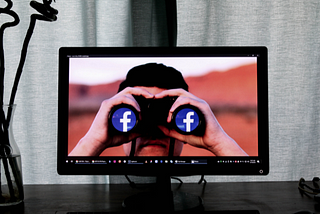 A desktop computer screen on a desk displaying an image of a person facing the viewer holding binoculars that have Facebook icons in each of the binocular lenses as if they were looking at Facebook through the binoculars.