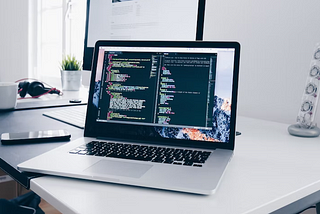 A laptop on a white desk and its screen show some code written in code editor software.