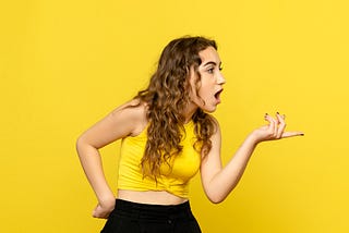 Young woman against a bright yellow background, pointing to someone out of view and looking shocked.