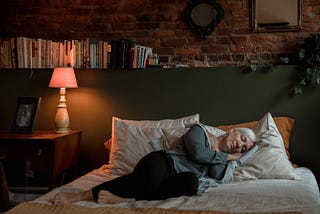 An older woman with grey hair sleeping on top of her bed with all her clothes on in a dimly lit room.