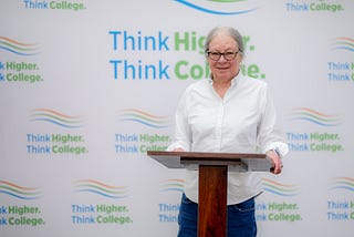 Debra Hart, a white woman with long grey hair and glasses, smiles at the camera from a podium in front of a “Think Higher. Think College.” background.