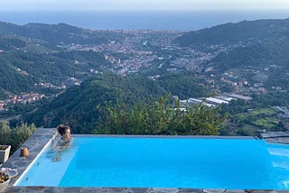 Woman sitting in a pool overlooking a town in Italy. Ocean is visible beyond.