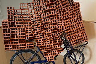 Dozens of large red clay mud bricks loaded on a black metal bicycle in an art installation of 21st century art. The bricks seemingly overwhelm the bike, which is still, however, standing.