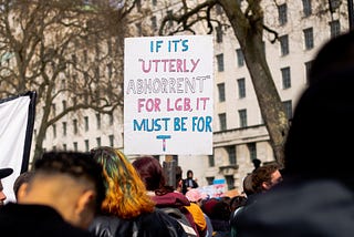 Photograph of a sign at Ban conversion therapy for all — The Trans Rights Protest London, April 2022 that reads “If it’s utterly abhorrent for LGB, it must be for T” in the transgender flag colors.
