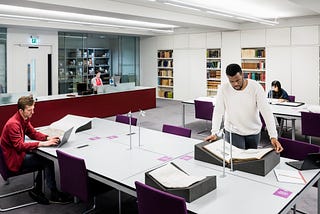 Four people are consulting archives and manuscripts in a library reading room. Three are sitting at desks and the fourth is standing to read a large volume.