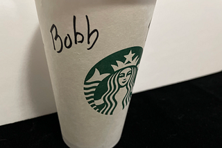 A Starbucks cup sits on a black counter. The name Bob (misspelled as “Bobb”) is written on it in black marker.