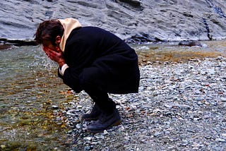 A man hunched over looking at water with his hands cupping his face.