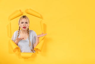 Grumpy looking woman, long blonde hair in plaits, red lipstick, pushing through yellow paper. Her hands are palm-up and pointing to her left as if she is explaining something to an idiot.
