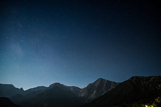 A night time scene with the stars in the sky over a mountain range. In the distance, there is light illuminating the horizon.