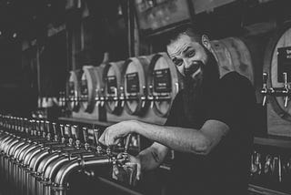 A person at a pub bar fills a pint glass, looking at the camera. There are several casks in the background.