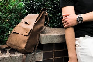 Brown bag on a ledge of a short brick wall next to a person holding their arm with a watch on.