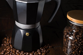 A black Moka pot coffee maker around coffee beans in a jar.