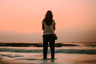 A woman stands alone at the edge of the ocean at sunrise, looking out across the water beneath a soft pink sky, symbolizing reflection, resilience, and new beginnings.