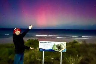 The author wearing a red hat standing next to a sign. Behind him, the ocean waves are visible with a bright red aurora in the night sky.