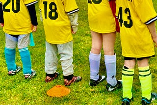 The backs of four children are in view. They are wearing soccer shirts and cleats.