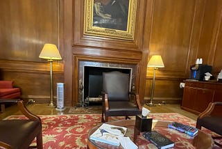 The inside of the Member Lounge at the American Museum of Natural History. Wood-panelled walls with a grand portrait above a fireplace and antique furniture on a beautiful red, floral carpet.
