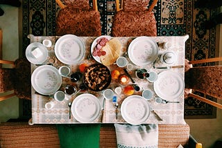 A colorful dinner setup with the meal on the white table and brown chairs with a huge cushioned bench and throw pillows.