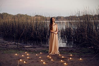 A woman in a white dress stands on the muddy bank of a lake. Lit candles are at her feet. The flora in the background hints the picture was taken in late fall. | Photo courtesy: Carolina Basi, Pexels