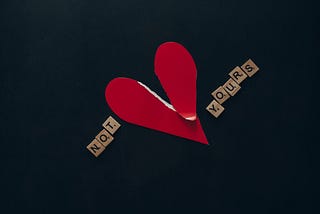 A red paper heart is torn in two and placed in the middle of wooden tiles which spell out the words “not yours”