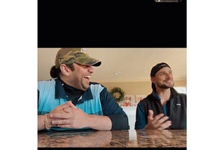 Two men (Pierre and Zach) are seated at a kitchen counter, sharing a jovial moment. Pierre is wearing a camouflaged baseball cap and a smile that reaches his eyes, his body language open and animated as he laughs. He has short, dark hair and is dressed in a blue and black striped golf shirt. Zach, also laughing, gestures with his hands as if emphasizing a point in a story. He wears a dark cap backwards with a logo at the front, and a black zip-up jacket.